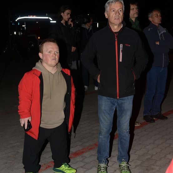 Ein Mann in einer schwarzen Jacke mit rotem Streifen steht neben einem Mann in einer roten Jacke, beide sehen ernst aus, mit anderen im Hintergrund bei Nacht.