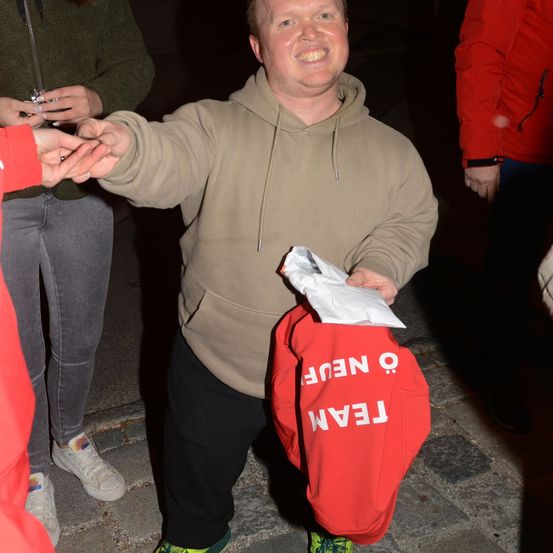 Ein Mann in einer beigen Kapuzenjacke hält eine rote Tasche mit dem Wort 'FEEL' darauf. Er lächelt und scheint sich in einer sozialen Umgebung zu befinden.