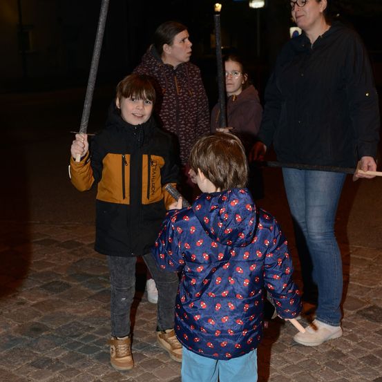 Eine Gruppe von Kindern und Erwachsenen steht nachts auf einer Kopfsteinpflasterstraße, hält Stöcke und lächelt. Die Kinder tragen Jacken mit Kapuzen, eines davon hat ein Spider-Man-Design.