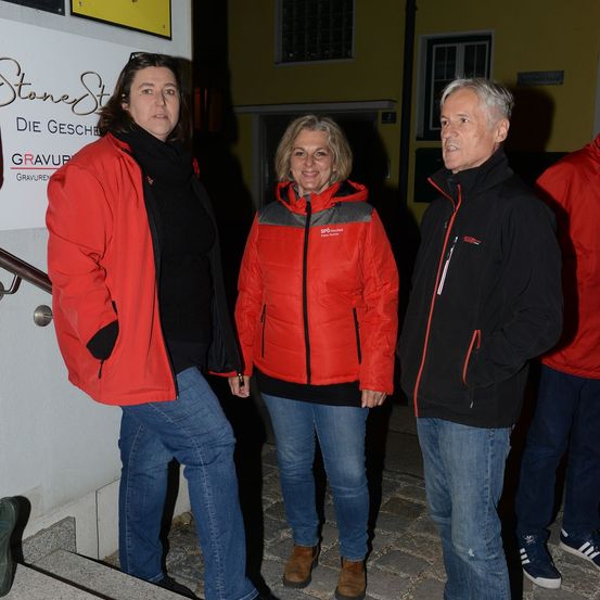 Drei Erwachsene, zwei Frauen und ein Mann, stehen nachts vor einem Gebäude. Sie sind in Wintermänteln gekleidet und scheinen zu plaudern. Die Frau auf der linken Seite trägt einen roten Mantel, während die Frau in der Mitte einen roten und grauen Mantel trägt. Der Mann auf der rechten Seite trägt einen schwarzen Mantel.