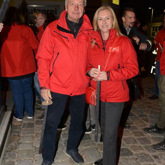 Ein älterer Mann und eine Frau in passenden roten Jacken stehen zusammen, lächeln und halten ein kleines Objekt in den Händen. Sie sind von anderen Menschen in roten Jacken umgeben, möglicherweise bei einer sozialen Veranstaltung.