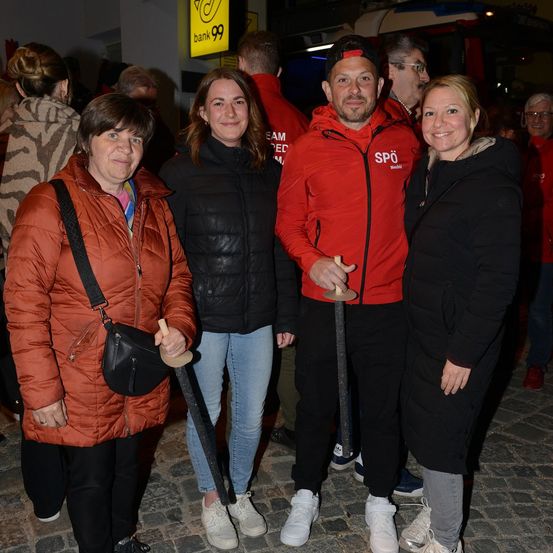 Vier Menschen stehen zusammen in einer Gruppe und lächeln für ein Foto. Sie sind in Winterkleidung gekleidet, wobei zwei Frauen Holzschwerter halten. Der Mann in der Mitte trägt eine rote Jacke mit den Buchstaben 'SPO' darauf. Hinter ihnen stehen weitere Menschen, und ein gelbes Schild mit der Nummer '99' ist sichtbar.
