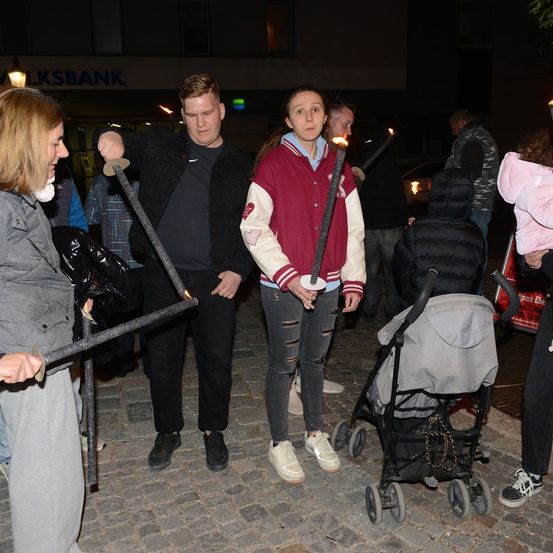 Eine Gruppe von Menschen mit Fackeln und in Winterkleidung steht nachts auf einer Kopfsteinpflasterstraße. Eine Frau in einer bordeauxroten Jacke hält eine Fackel, während ein Mann in einer schwarzen Jacke ein Schwert hält. Ein Kinderwagen steht in der Nähe, und ein Gebäude mit einem Schild, das 'WALKSBANK' liest, ist im Hintergrund.