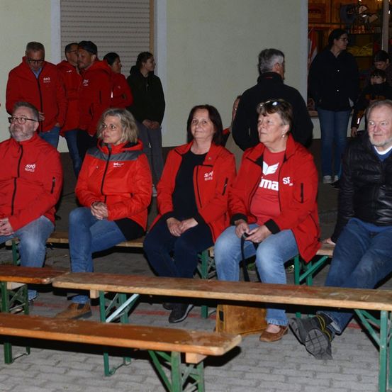 Eine Gruppe von Erwachsenen in roten Jacken sitzt auf Bänken, einige Leute stehen hinter ihnen. Sie befinden sich in einem Raum mit einem Ziegelboden und einer weißen Wand.