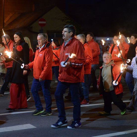 Eine Gruppe von Menschen in roten Jacken und mit brennenden Fackeln in der Hand gehen nachts in einer Reihe auf einer Straße.
