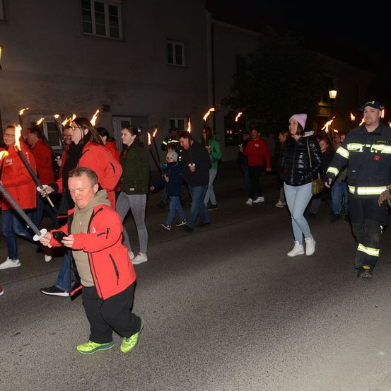 Eine Gruppe von Menschen, die in Rot und Schwarz gekleidet sind, marschiert bei einer nächtlichen Parade und hält brennende Fackeln. Ein Mann vorne beugt sich nach unten und hält eine Fackel.