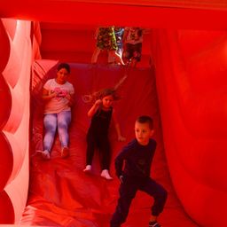 Drei Kinder rutschen einen roten aufblasbaren Rutschvergnügen hinunter, eine Mädchen in Weiß und eine in Schwarz. Ein Junge in Blau beobachtet vom Boden.