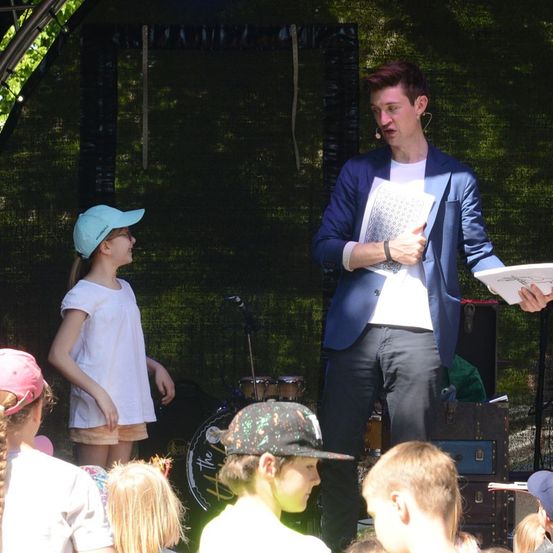 Ein Mann in einem blauen Anzug hält ein Buch in der Hand und spricht zu einer Gruppe von Kindern auf der Bühne. Ein Mädchen in einem weißen Shirt und ein Junge in einer Mütze hören aufmerksam zu.