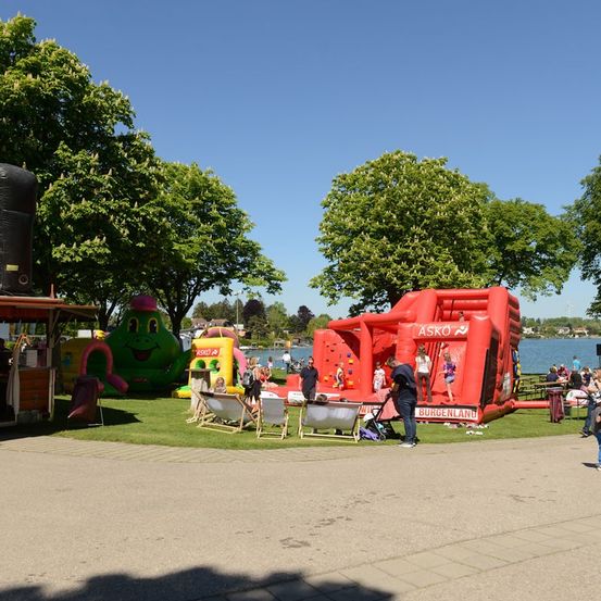 Ein Park mit See, aufblasbarem Spielplatz und Menschen, die den Tag genießen. Bäume und ein Imbissstand sind ebenfalls sichtbar.