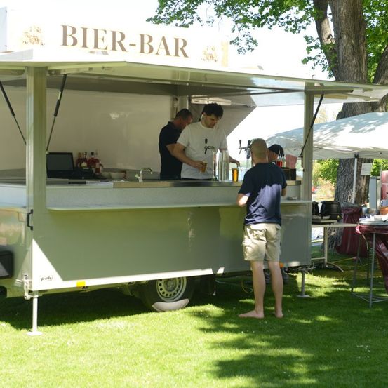 Ein mobiler Bierstand mit zwei Barkeepern, die einem Kunden in Shorts an einem sonnigen Tag Getränke servieren.