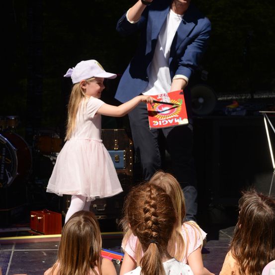Ein Zauberer auf der Bühne mit einem kleinen Mädchen, das eine rote Box mit der Aufschrift 'Magie' hält, während andere Kinder zusehen.