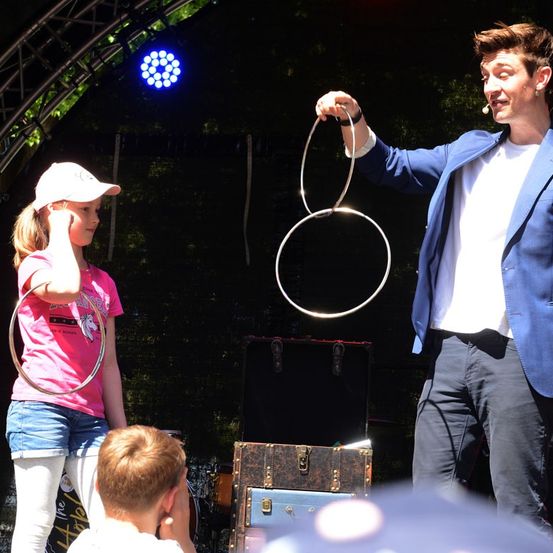 Ein Mann führt mit einem jungen Mädchen auf der Bühne einen Hula-Hoop-Trick durch, während ein Junge vorne zusieht.