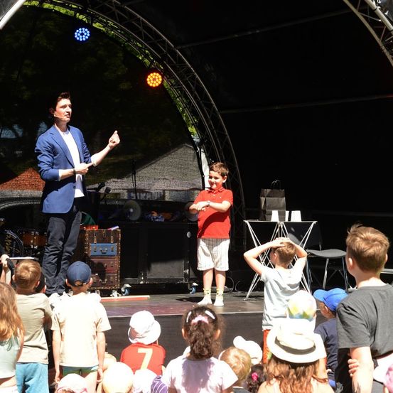 Ein Zauberer in einem blauen Anzug führt mit einem jungen Jungen in einem roten Shirt auf der Bühne vor, umgeben von einer Menge von Kindern.