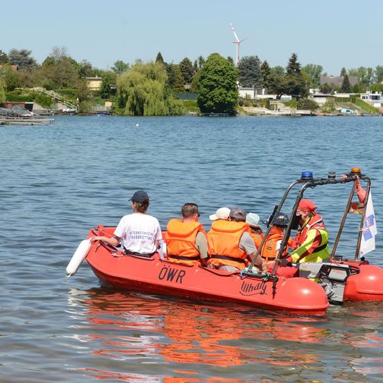 Fünf Personen in Rettungswesten fahren in einem roten Boot auf einem See mit einer Flagge hinten. Bäume und eine Windturbine sind im Hintergrund sichtbar.