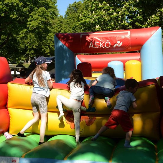 Vier Kinder spielen auf einem bunten aufblasbaren Sprunghaus mit dem Wort Askö darauf. Die Kinder tragen Freizeitkleidung und scheinen viel Spaß zu haben.