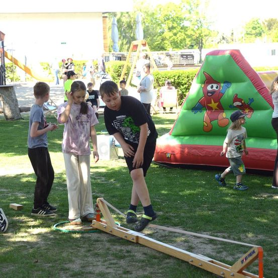 Eine Gruppe von Kindern spielt auf einem Rasenfeld mit einem hölzernen Wippgerät und einem bunten aufblasbaren Schloss im Hintergrund.