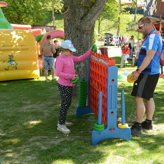 Ein junges Mädchen spielt mit einem Mann ein Riesen-Spiel von Connect Four, während andere im Hintergrund in der Nähe von Luftballons und einem Baum sind.