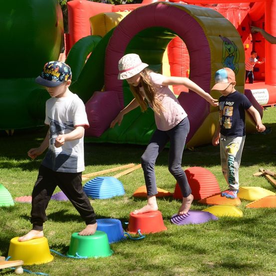 Drei Kinder spielen auf einem Feld mit bunten Bechern, ein Mädchen balanciert auf einem roten Becher, während zwei Jungen zusehen. Hinter ihnen sind aufblasbare Spielgeräte aufgebaut.