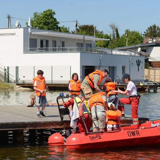 Eine Gruppe von Menschen in orangefarbenen Rettungswesten besteigt ein rotes Boot an einem Steg. Ein Gebäude mit einer Flagge ist im Hintergrund zu sehen.