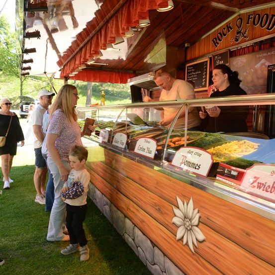Eine Familie steht in einer Schlange vor einem Imbisswagen mit Holzverkleidung, beschriftet mit 'Rock & Food.' Das Menü umfasst Artikel wie 'star crisp pommes' und 'Zwiebeln.' Der Verkäufer serviert Speisen durch eine Glasvitrine.