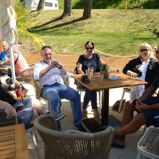 Eine Gruppe von Menschen sitzt um einen Tisch in einer Außenumgebung, einige tragen Sonnenbrillen und lächeln. Auf dem Tisch steht eine Ananas, und im Hintergrund befindet sich ein Rasen mit Bäumen.