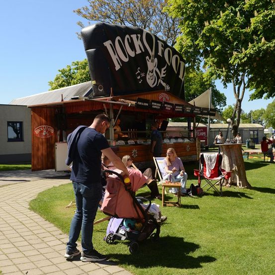Ein Mann schiebt einen Kinderwagen mit einem Baby an einem sonnigen Tag in einem Park in der Nähe eines Essenswagens mit der Aufschrift 'ROCKY FOOD'.