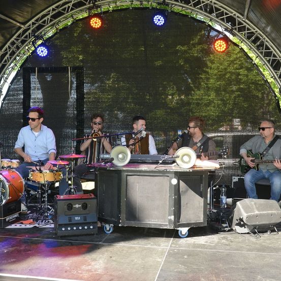 Fünf Musiker spielen auf der Bühne mit einem Schlagzeug, Trompete, Gitarre und Mikrofonen. Die Bühne ist von bunten Bühnenlichtern beleuchtet.