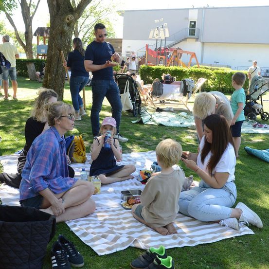 Eine Gruppe von Menschen macht ein Picknick auf dem Rasen in einem Park, ein Mann steht und hält Essen.