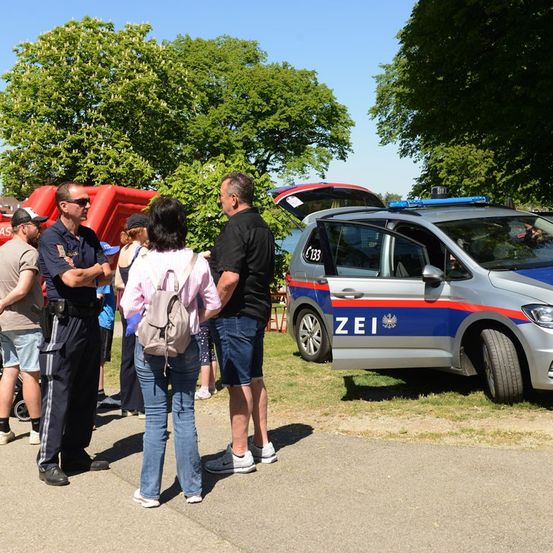 Eine Gruppe von Menschen steht in der Nähe eines Polizeiwagens mit offener Tür. Ein Polizist spricht mit einer Frau mit Rucksack.