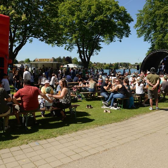 Eine große Menschenmenge versammelt sich in einem Außenbereich, sitzt auf Bänken und Tischen, wahrscheinlich für ein Festival. Im Hintergrund befinden sich Bäume, eine Bühne und ein Gewässer.