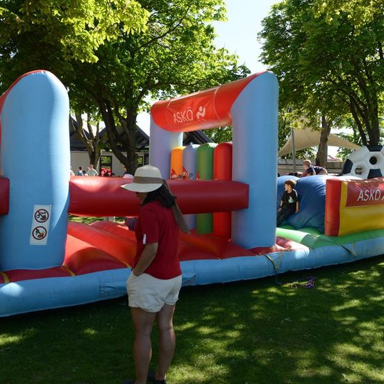 Eine Frau in einem roten T-Shirt und weißen Shorts steht neben einem großen aufblasbaren Hindernisparcours, in dem Kinder spielen. Das aufblasbare Objekt ist bunt und mit 'ASKO' gekennzeichnet. Bäume und ein Gebäude sind im Hintergrund zu sehen.