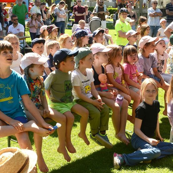 Eine Gruppe von Kindern und Erwachsenen versammelt sich auf einem Rasenfeld, einige sitzen auf Bänken und andere stehen. Die Kinder tragen Mützen und Turnschuhe, und einige halten Spielzeug. Die Erwachsenen stehen hinter den Kindern, einige mit verschränkten Armen. In der Ferne sind weitere Menschen und ein weißes Zelt zu sehen.