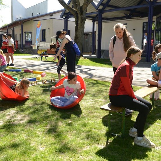 Mehrere Kinder spielen in einem Spielplatzbereich, während Erwachsene beaufsichtigen. Ein Mädchen sitzt auf einem roten Beanbag-Stuhl, während ein anderes Mädchen auf einer Bank sitzt.
