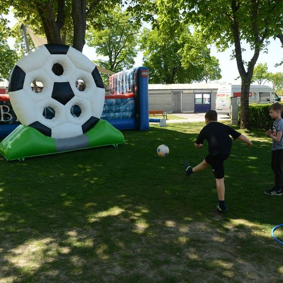 Zwei Jungen spielen Fußball auf einem Rasenfeld mit einem großen aufblasbaren Fußball im Hintergrund.