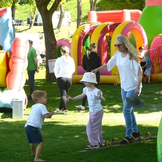 Eine Frau führt ein junges Mädchen und einen Jungen auf einem Balancebalken in einem Park mit bunten Luftschlössern im Hintergrund.