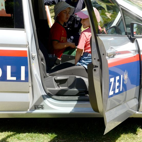 Zwei Kinder, eines in einem roten Shirt und das andere in einem rosa Shirt, sitzen in einem offenen Polizeiwagen mit offener Tür. Das Auto hat das Wort 'POLI' an der Seite und 'ZEI' an der Tür.