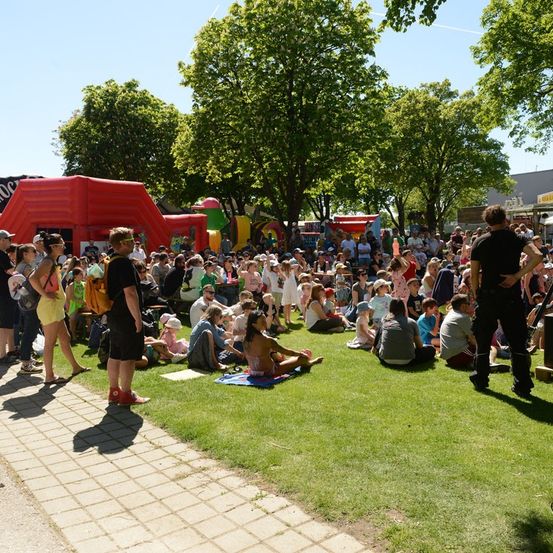 Eine große Menschenmenge, darunter auch Kinder, versammelt sich in einem Park mit aufblasbaren Spielgeräten und Bäumen im Hintergrund.