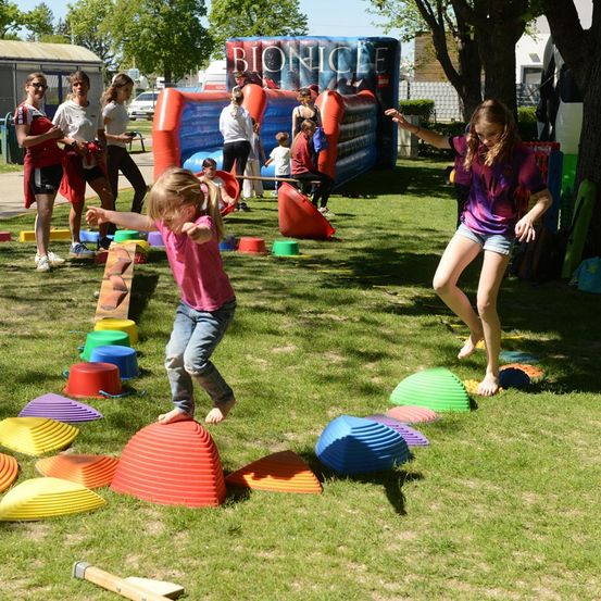 Kinder spielen auf einem Rasenfeld mit verschiedenen farbigen Hindernissen, einschließlich Kegel und einem Hammer. Ein Mädchen in einem rosa Shirt springt über einen roten Kegel. Im Hintergrund befindet sich ein aufblasbares Hüpfhaus mit der Aufschrift 'Bionicle'.