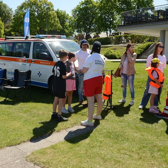 Eine Gruppe von Menschen, darunter Kinder, steht an einem sonnigen Tag vor einem Krankenwagen. Sie tragen Rettungswesten und scheinen ein Gespräch zu führen.