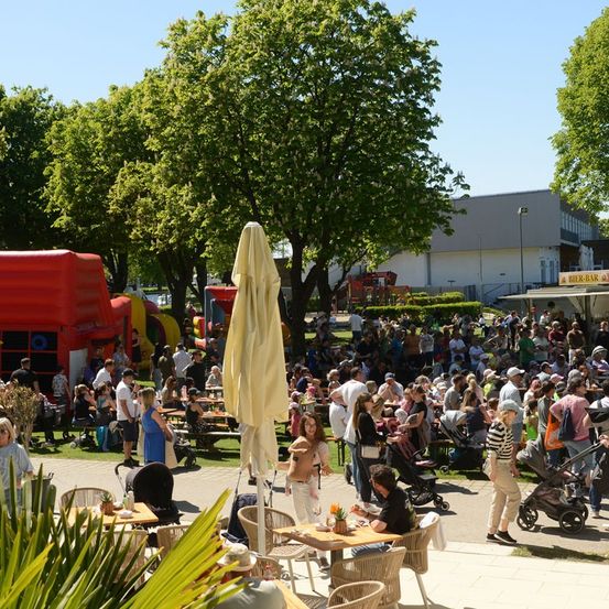 Eine große Menschenmenge versammelt sich in einem Außenpark und genießt Essen und Getränke an Tischen unter Sonnenschirmen. Es gibt aufblasbare Spielplätze und ein Gebäude mit einem Schild, das Bier-Bar liest.