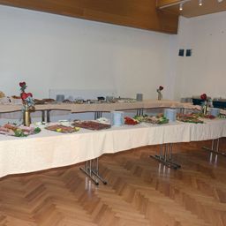 Ein langer Tisch, der mit einer weißen Tischdecke bedeckt ist, ist mit verschiedenen Speisen wie Fleisch, Gemüse und Brot sowie Tellern, Schüsseln und Blumen in Vasen gedeckt.