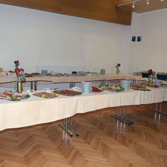 Ein langer Tisch, der mit einer weißen Tischdecke bedeckt ist, ist mit verschiedenen Speisen wie Fleisch, Gemüse und Brot sowie Tellern, Schüsseln und Blumen in Vasen gedeckt.