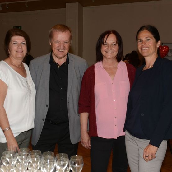 Vier Erwachsene, zwei Männer und zwei Frauen, lächeln und posieren für ein Foto vor einem Tisch mit Weingläsern.