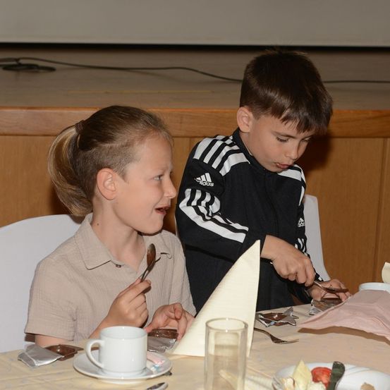 Zwei Kinder sitzen an einem Tisch, eines hält einen Löffel und das andere eine Gabel und beide schauen auf ihr Essen. Der Tisch ist mit Tellern, Tassen und Servietten gedeckt. Der Hintergrund ist unscharf, was auf eine Innenumgebung hindeutet.