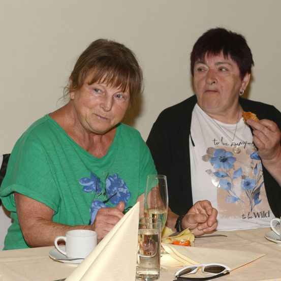 Zwei Frauen sitzen an einem Tisch, eine trägt ein grünes Shirt mit blauem Blumendesign, die andere ein weißes Shirt mit Blumenprint, isst eine Kleinigkeit.
