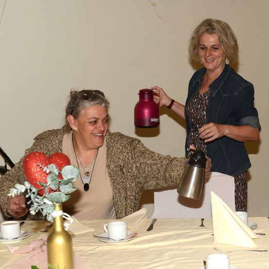 Zwei Frauen sitzen an einem Tisch, eine hält einen roten Thermos und die andere einen silbernen Thermos. Der Tisch ist mit Tassen, Untertassen und einer Blumenarrangement gedeckt.