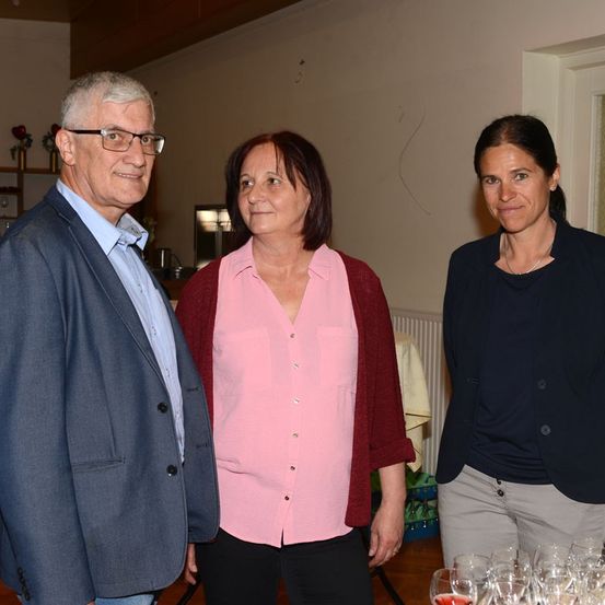 Drei Menschen stehen lächelnd in einem Raum. Ein Mann links, eine Frau in der Mitte in einem rosa Shirt und eine weitere Frau rechts in einer marineblauen Jacke. Hinter ihnen ist eine Wand mit einem Regal und einer weißen Tür.