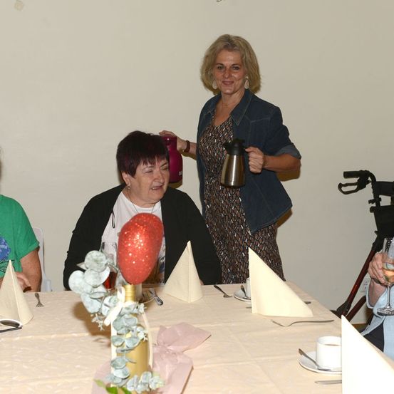 Eine Frau in einer Jeansjacke steht hinter einem Tisch mit einem roten Ballon als Mittelpunkt und lächelt, während sie eine Kaffeekanne hält. Eine andere Frau sitzt am Tisch und lacht.