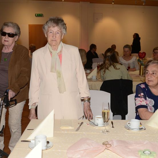 Drei ältere Frauen sitzen an einem Tisch in einem Essbereich. Eine Frau steht, trägt einen weißen Blazer und eine Krawatte. Eine andere Frau hält einen Stock.