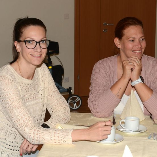 Zwei Frauen sitzen an einem Tisch, eine mit Brille und Halskette, die andere mit einer Uhr, beide lächeln und halten Tassen.